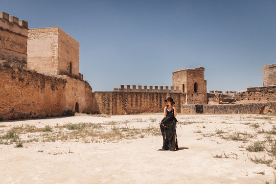 Stylish woman in black hat and long dress standing inside walls of old abandoned fortress on sunny day