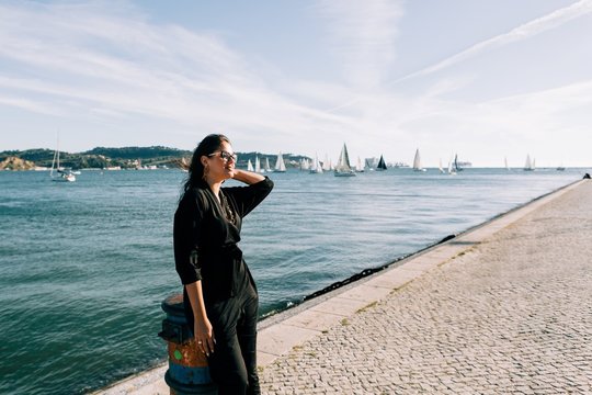 Side view of young attractive woman in black outfit standing by sea and leaning on pole in Lisbon