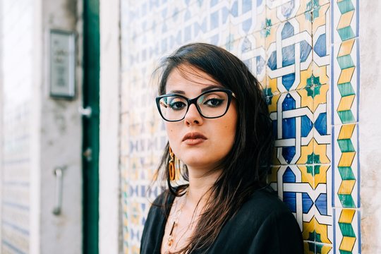 Attractive young woman in glasses and black blouse standing by colorful vintage wall and looking at camera in Lisbon