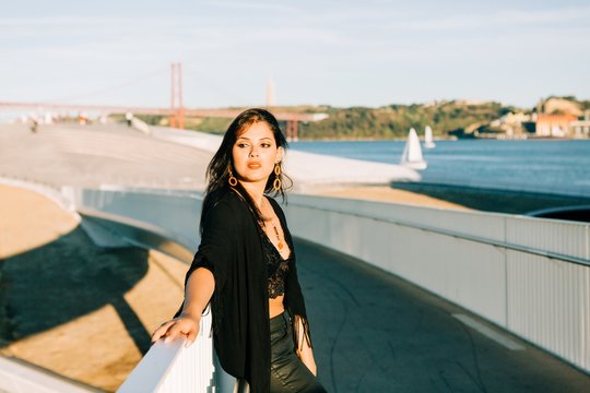Beautiful stylish woman in black outfit standing by bridge with city landscape in Lisbon on sunny day