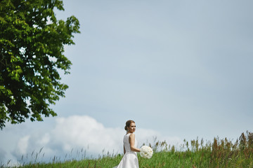 Beautiful young blonde woman in a stylish white dress with wedding hairstyle keeping a bouquet of white flowers and posing outdoors in a green field. Young bride walking before the wedding ceremony