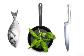 Fresh fish dorado, knife and frying pan with basil leaves on a white background. View from above.