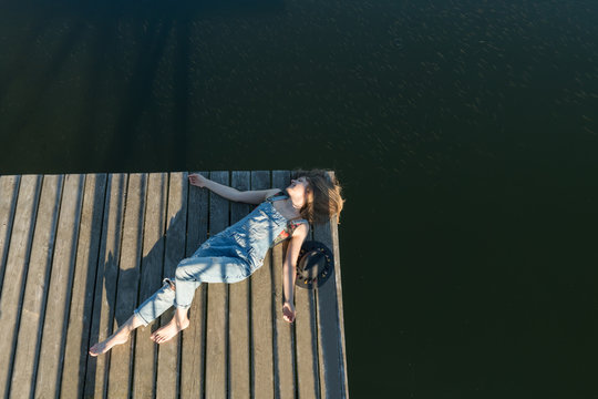 Top view of beautiful woman in black swimsuit and hat lying down on wooden pier of lake on clear blue sky and forest background