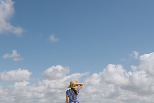 Content adult woman with blowing hair and in sundress walking with straw hat in hand on cloudy sky background
