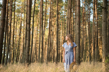 Happy adult woman in straw hat and sundress standing in forest among coniferous trees in golden sun ray smiling away