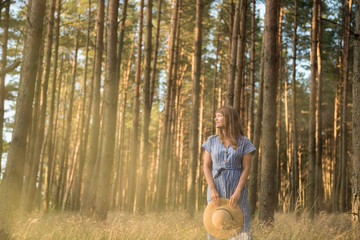 Happy adult woman in straw hat and sundress standing in forest among coniferous trees in golden sun ray smiling away