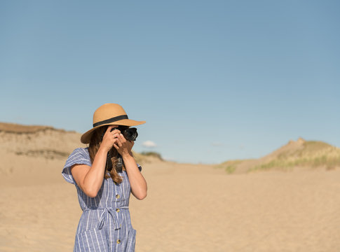 adult woman in straw hat and dress with camera taking photo on sand dune of beach in a sunny day