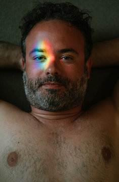 From above adult bearded naked man with hands behind head and rainbow light on eye lying in shadow