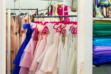 Rack with many beautiful holiday dresses for girls on hangers at children fashion showroom indoor. Kid girl dress hire or sewing studio for celebration birthday party or photography session event