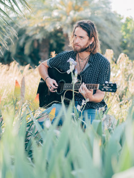 Pensive bearded hipster man sitting in meadow playing guitar looking away
