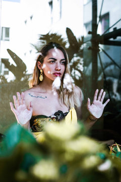 Young pretty serious confident female with bright make-up and tattoo looking away and touching glass with hands on light background