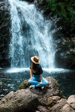 Cheerful female traveler in hat smiling and looking at camera while sitting on wet boulder near waterfall
