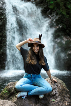 Cheerful female traveler in hat smiling and looking at camera while sitting on wet boulder near waterfall