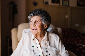 Portrait of happy senior curly gray haired woman in white shirt and with beads on neck looking away at home