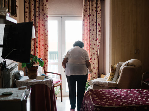 Back view of unrecognizable elderly woman walking to window in bedroom