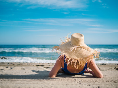Back view of pretty woman in hat with fringe at edge and dark blue swimsuit on sandy seaside looking at foam waves under turquoise cloudy sky