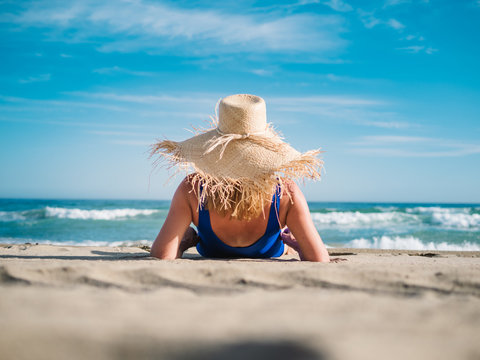 Back view of pretty woman in hat with fringe at edge and dark blue swimsuit on sandy seaside looking at foam waves under turquoise cloudy sky