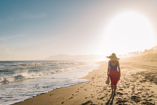 Back view of slim woman in hat and pareo walking along coastline in bright sunlight