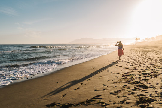 Back view of slim woman in hat and pareo walking along coastline in bright sunlight