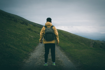 Back view of unrecognizable male with backpack standing on rough path on grassy hill slope against gray overcast sky in nature