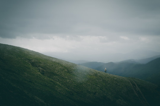 Back view of unrecognizable male with backpack walking on rough path on grassy hill slope against gray overcast sky in nature