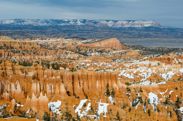 Sun lit Bryce Canyon in detailed (still broad) view. Red / ochre canyon rock formations in places overgrown by green pines, partly covered by white snow. Mountains on distant horizon