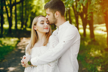 Fototapeta premium A beautiful and gentle girl with light hair and a white dress is walking in a sunny summer forest with her handsome guy in a white shirt and dark pants