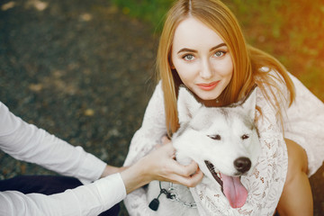 A beautiful and gentle girl with light hair and a white dress is walking in a sunny summer forest with her handsome guy in a white shirt and dark pants and they playing with cute gray dog