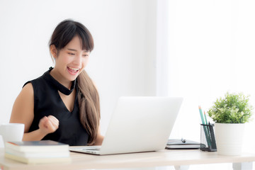 Beautiful portrait asian young woman smile working online laptop computer with happy satisfied sitting on desk office, girl using notebook with excited and glad, business and success concept.