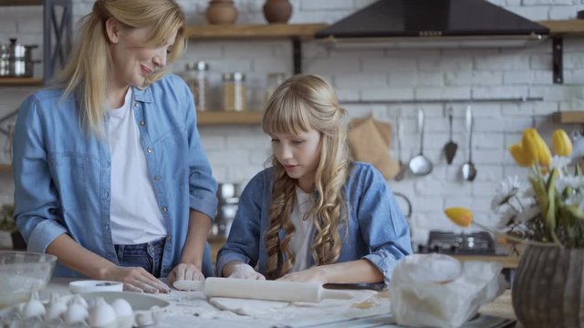 Mom and daughter bake pizza in the kitchen together. girl helps her mom to roll out the dough with a rolling pin. 4K