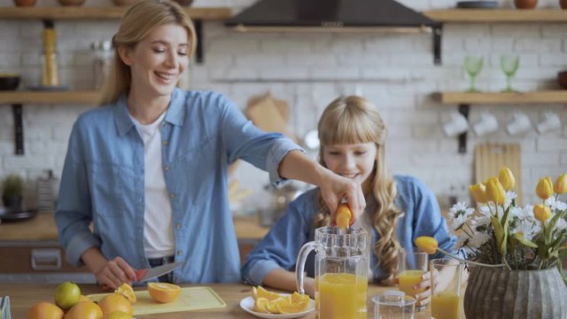 Mom and daughter make natural orange juice in the kitchen. Charming young mother squeezes juice from an orange into a jug. 4K