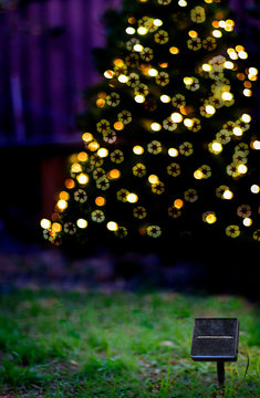 A solar panel in front of a Christmas tree with solar powered Christmas lights at dusk with recycle symbol bokeh. Sustainable energy and living concept.