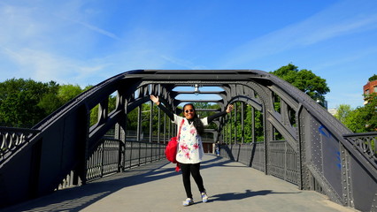 junge frau steht lächelnd auf alter brücke und hebt erfolgreich die Arme vor blauem Himmel