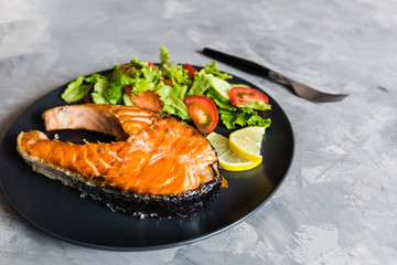 Fried salmon fish steak with vegetable salad and lemon on black plate. Creative layout made of fish, lettuce, tomato, cucumber. Healthy food concept