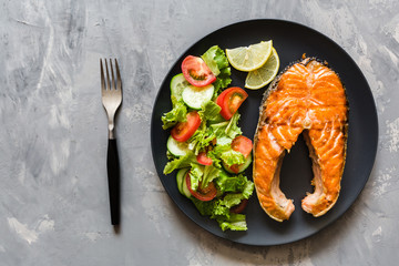 Fried salmon fish steak with vegetable salad and lemon on black plate. Creative layout made of fish, lettuce, tomato, cucumber, top view, flat lay, mockup, overhead. Healthy food concept