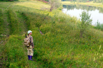 Yakut Asian girl tourist fisherman dressed in protective color clothes stands with a backpack and a fishing rod in his hand on the trail at the wild lake in the North of the taiga.