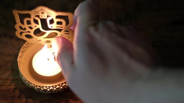Closeup of a man's hand lighting a wax tea candle in front of Lord Ganesha golden cutout during Diwali, festival oof lights, worship celebrations with a lighter in dark to show the light.