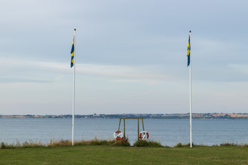 Fototapeta na wymiar Lifebuoy on the Swedish island of Ven in the southern parts of the country during a warm summer evening. Safety device below two Swedish flags next to a stone beach. 