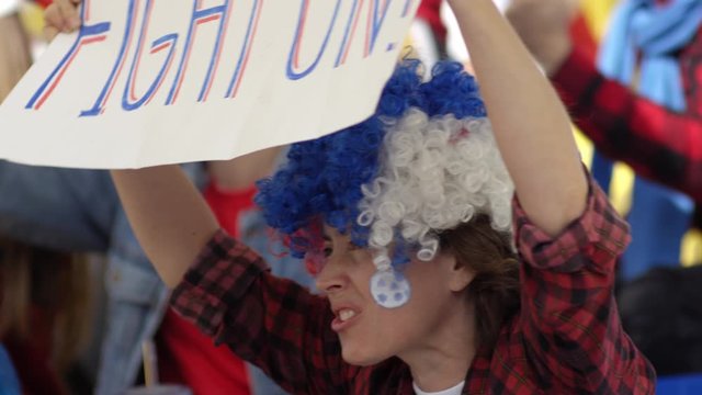 Chest-up shot of young Caucasian woman in colorful wig and plaid shirt sitting at sports match, shouting with excitement, waving banner and jumping up from seat, and man blowing vuvuzela in background