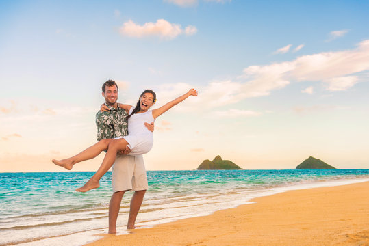 Happy honeymoon couple on beach wedding vacation newlyweds excited in Hawaii travel destination. Husband carrying Asian woman wife with arm up on Lanikai beach, Oahu, Hawaii.
