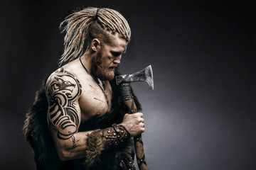 Medieval warrior berserk Viking with tattoo on skin, red beard and braids in hair with axe and shield attacks enemy. Concept historical photo - obrazy, fototapety, plakaty