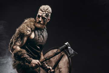 Medieval warrior berserk Viking with tattoo on skin, red beard and braids in hair with axe and shield attacks enemy. Concept historical photo - 285178992