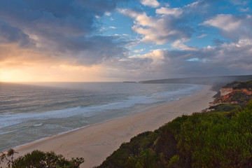 The sun rises and lights up the clouds with wonderful oranges and yellows, as the mist from the ocean spreads itself across the beach and bush areas on Terrace Beach in NSW Australia