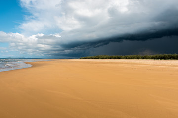 The sea water softs the bright orange sands as a thunderstorm bring dark dramatic clouds that loom over the lush and dense forests on Middle Island near 1770 in Australia.