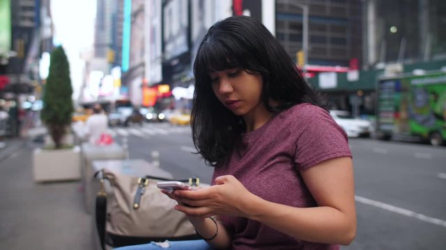 A young Latina woman sits in Times Sq, texting on her phone. Slow motion.