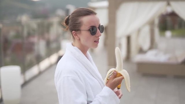Woman in robe peels and eats a banana on balcony, medium shot handheld
