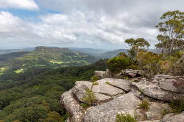 Fototapeta na wymiar A rocky and grey cliff edge with a tree on top, looks over the dense Australia forest with a mountain in the distance on a cloudy day at Drawing Room Rocks in NSW, Australia