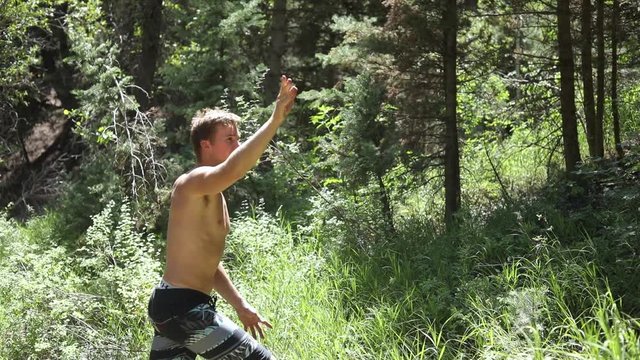 Slow motion shot of a shirtless man practicing his axe throwing skill by trying to throw his hatchet at a pine tree.