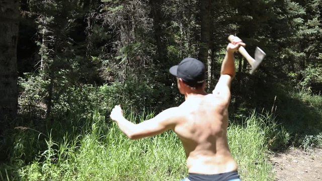 Slow motion shot of a shirtless man practicing his axe throwing skill by trying to throw his hatchet at a pine tree.