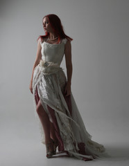 full length portrait of red haired girl wearing torn and tattered wedding dress. Standing pose against a studio background with contrasty shadow lighting.